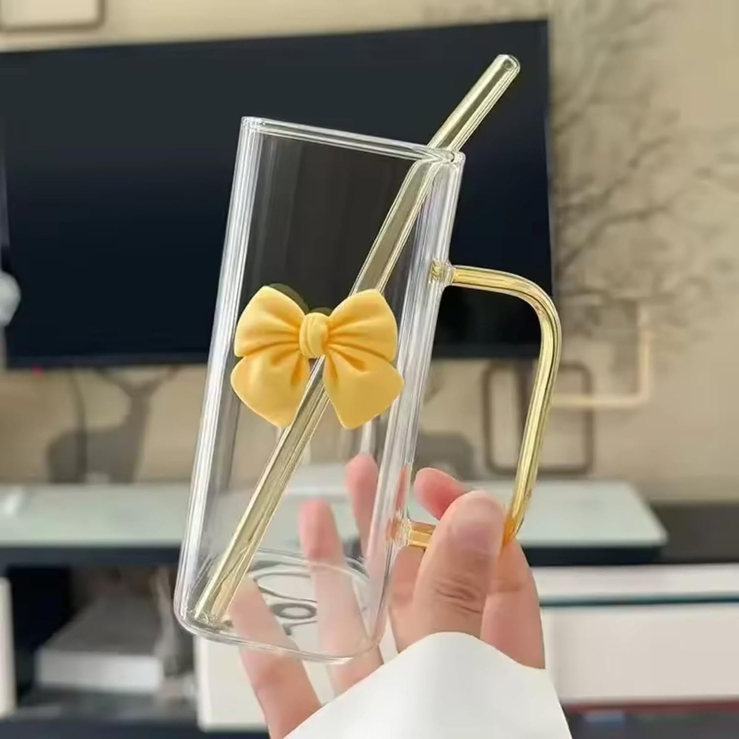 Clear glass mug with gold handle and yellow bow held by a hand in an indoor setting.