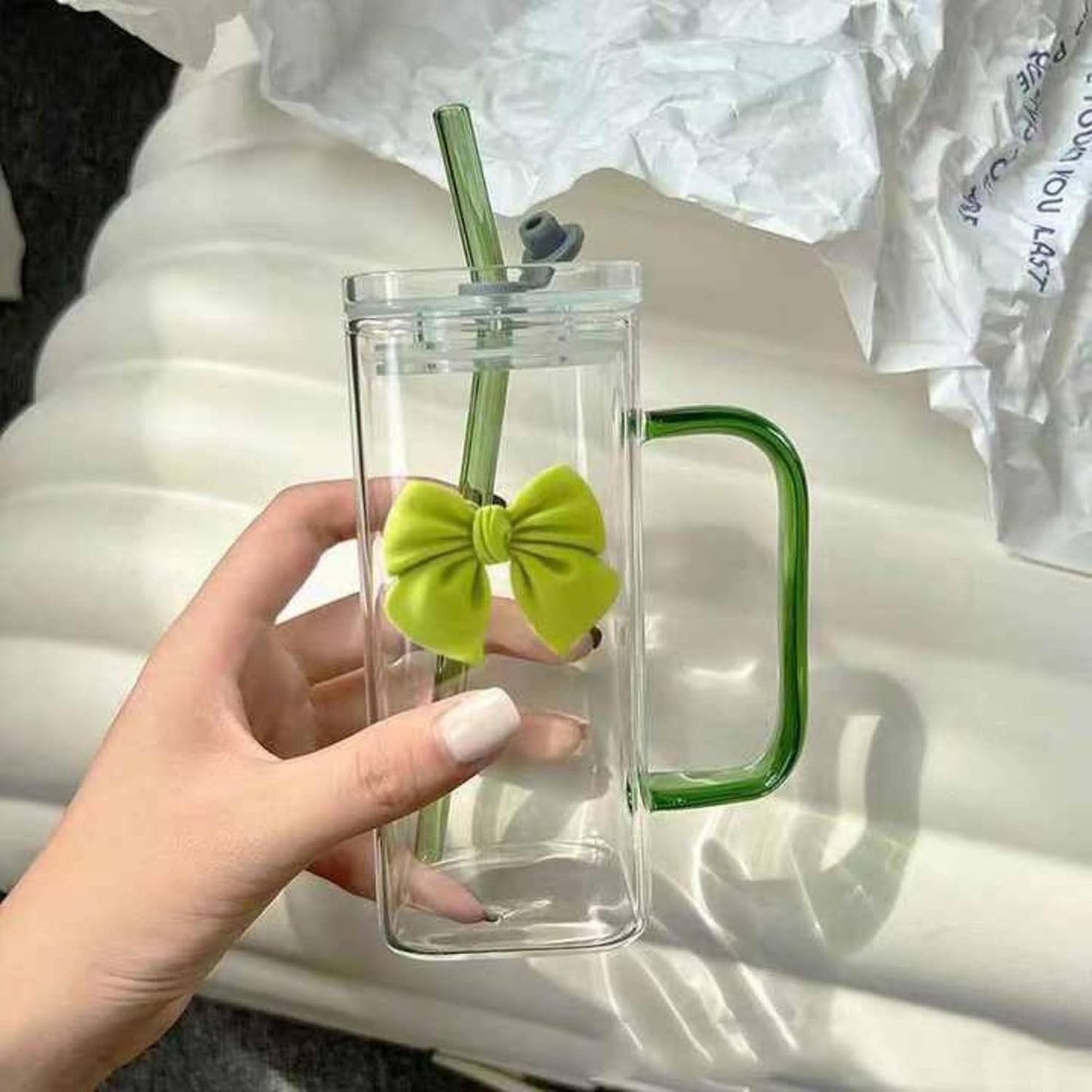 Clear mug with green handle and bow, held by a hand on a white surface.