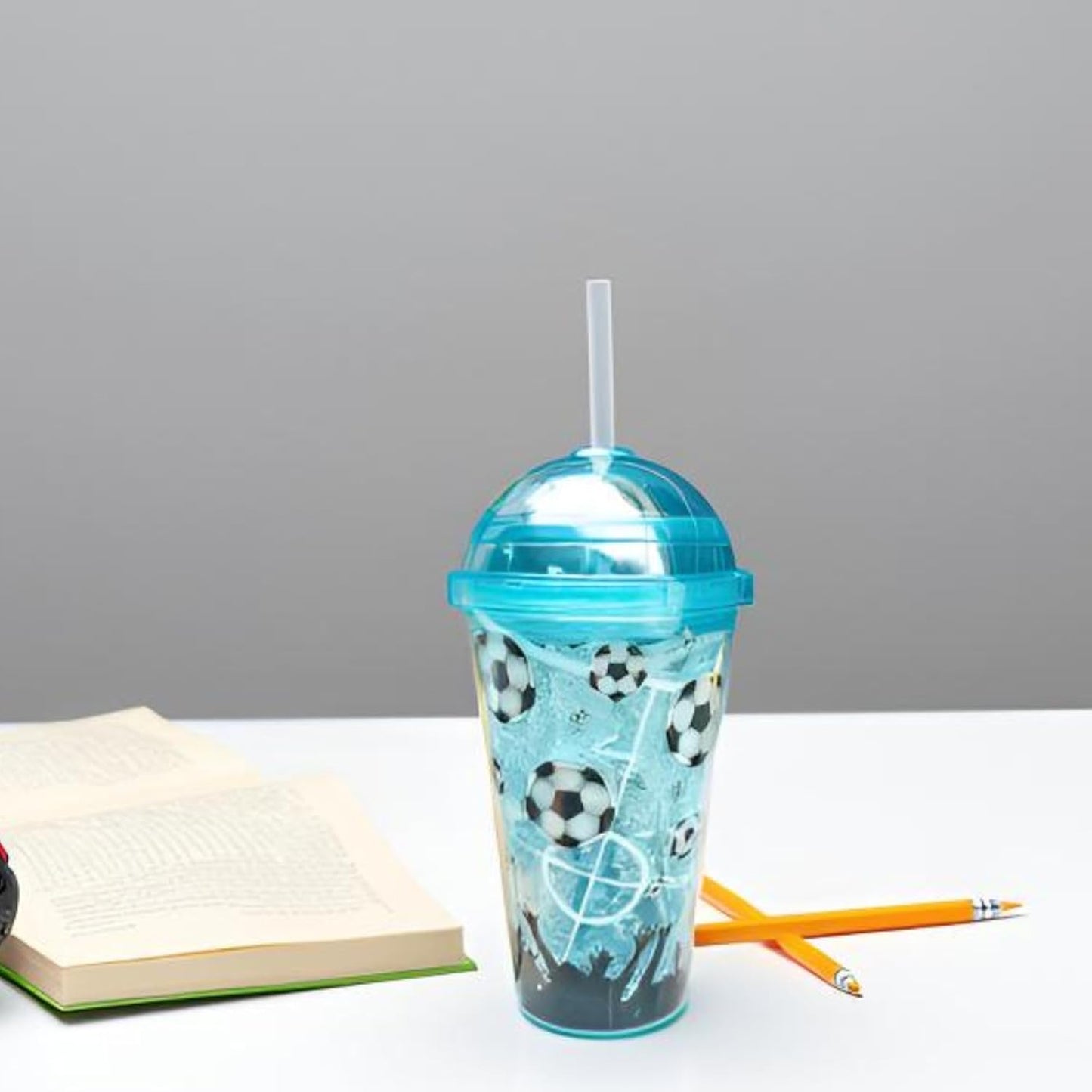 Clear tumbler with soccer ball design and blue lid on a white surface with an open book and pencils.