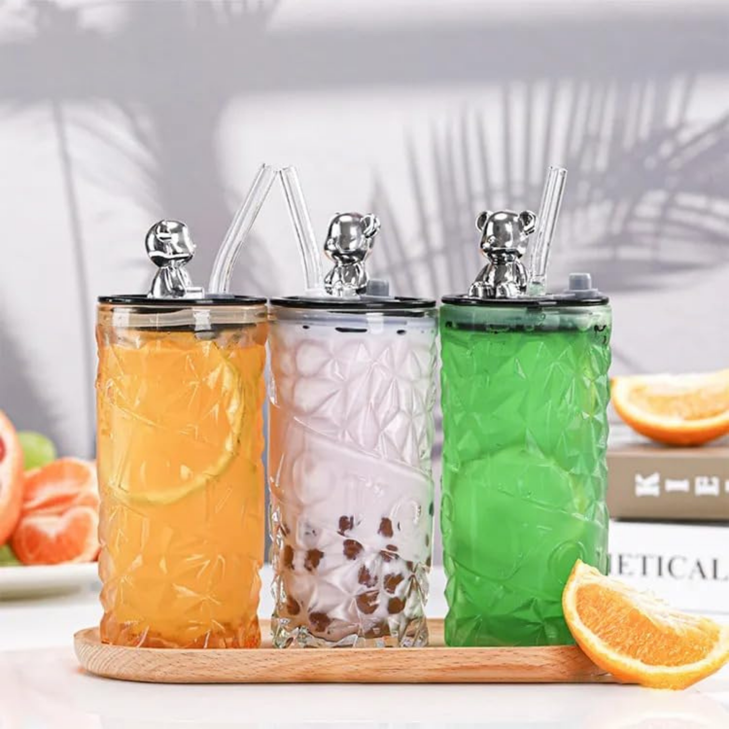 Three glass tumblers with lids and straws on a wooden tray, filled with orange, white, and green beverages.