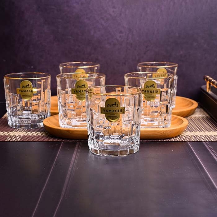 Five clear cut-glass rocks tumblers with gold TAMASIN labels, arranged on round wooden coasters against a dark purple textured background.