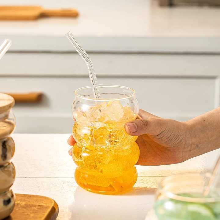 Hand holding a glass of iced yellow beverage with a straw on a kitchen counter.