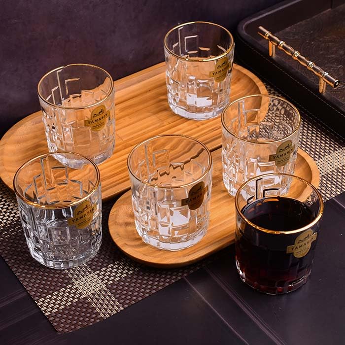 Five clear cut-glass rocks glasses with gold rims and gold labels, arranged on two wooden trays with one glass filled with dark red liquid, set on a dark table.