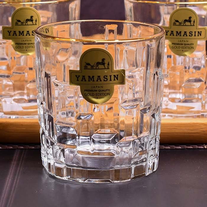 A clear faceted whiskey glass with a gold Yamasin label, shown in the foreground with other matching glasses blurred in the background on a wooden tray.