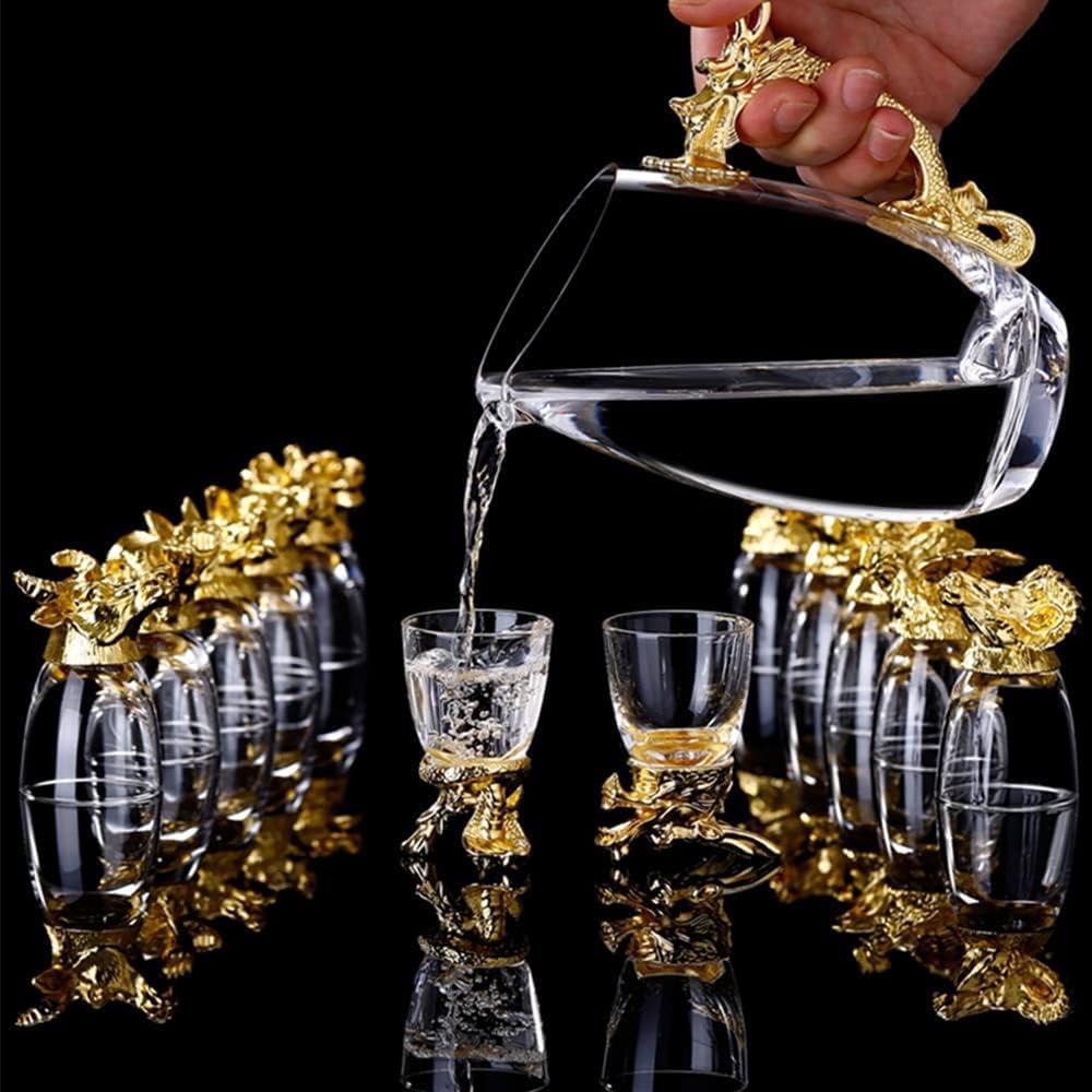 A hand holds a clear glass pitcher with ornate gold dragon-style handles and spout, pouring liquid into small matching clear glasses with gold bases, arranged on a glossy black reflective surface.