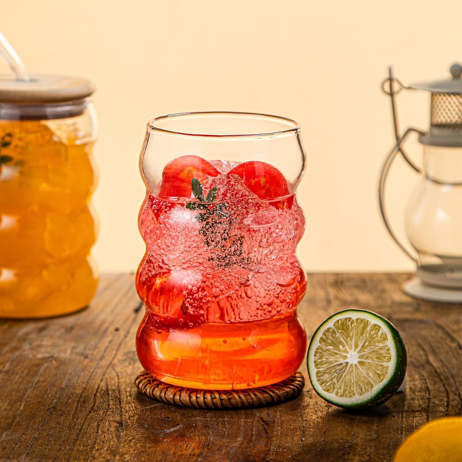 Glass with red drink and ice cubes on a wooden table with a lantern and jar in the background.
