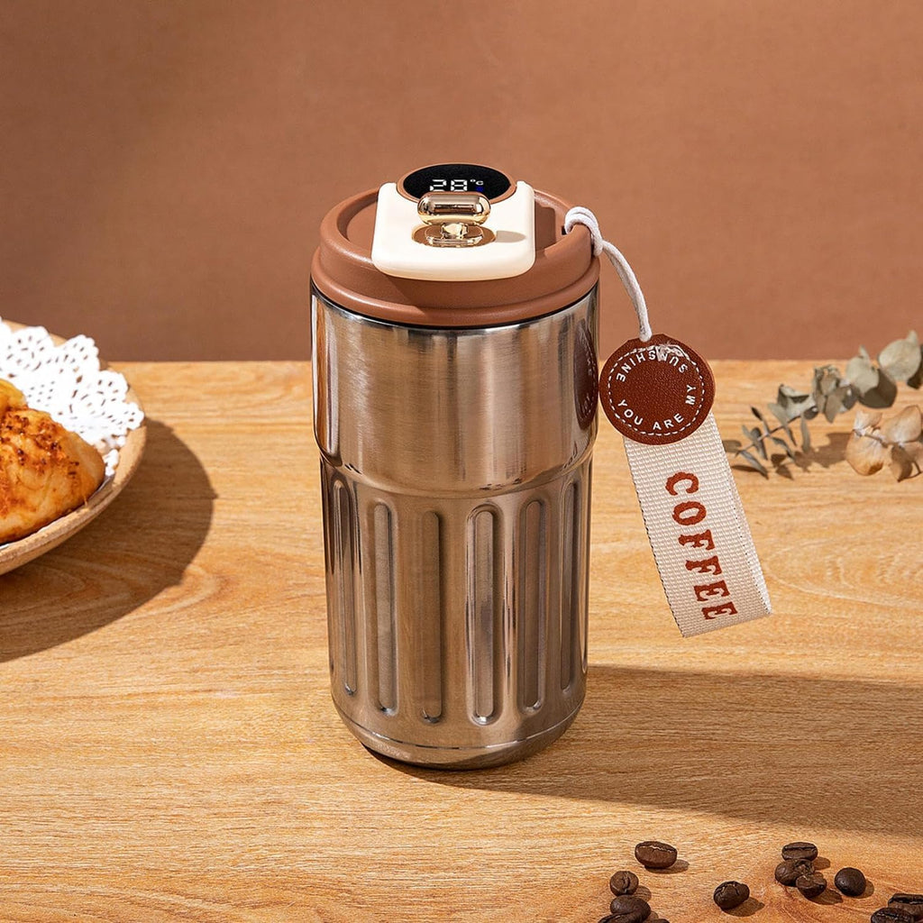 Stainless steel travel mug with brown lid on a wooden surface, surrounded by coffee beans and a tag.