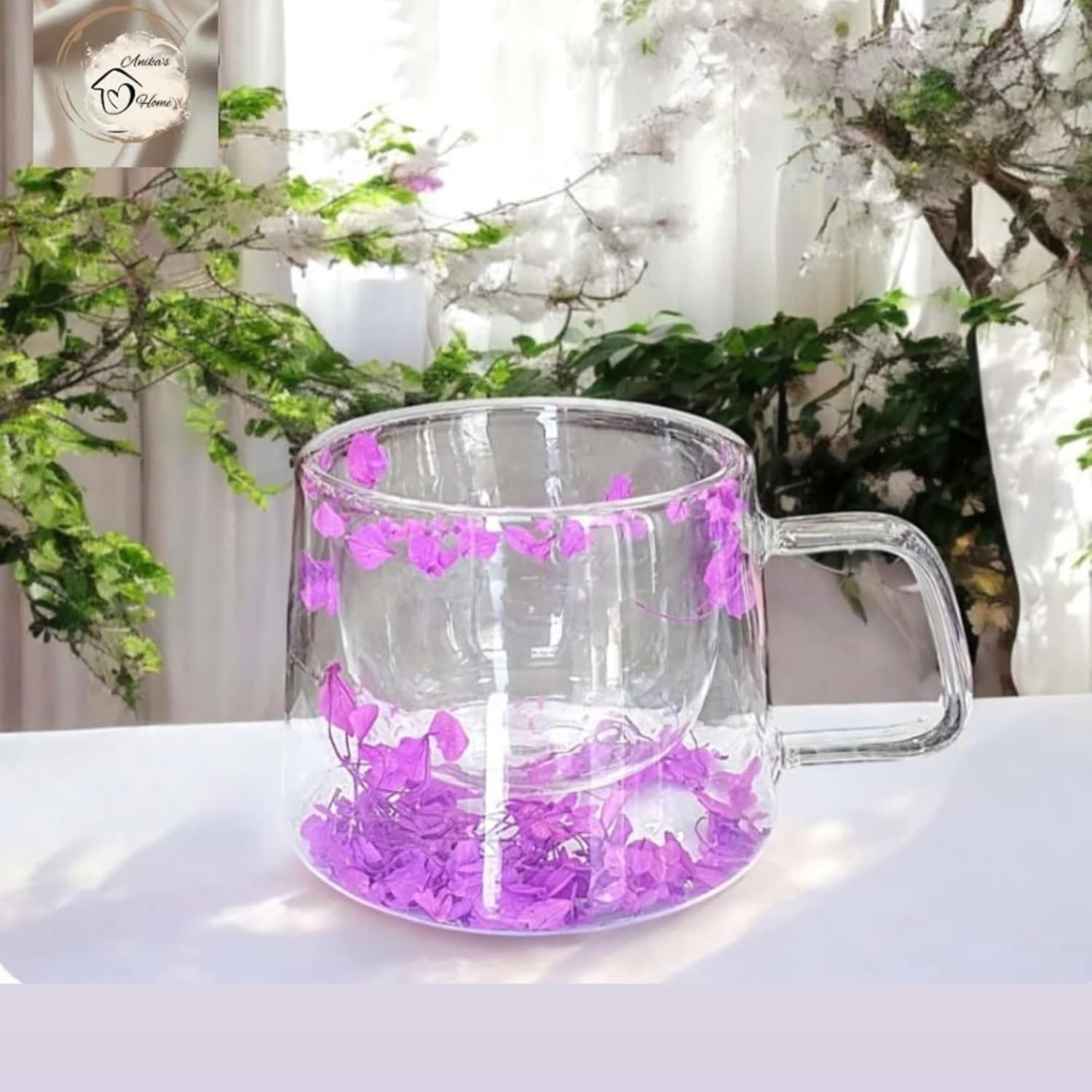 Clear glass mug with purple flower petals on a white surface with greenery in the background