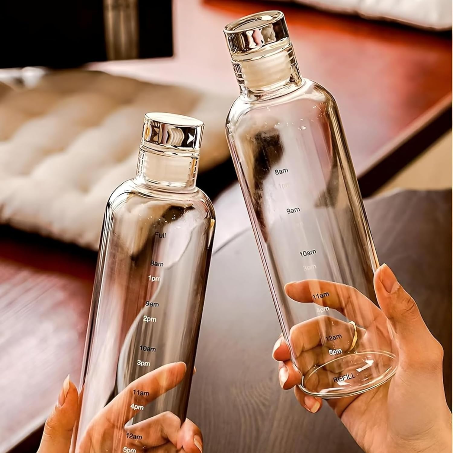 Two clear glass bottles with measurement markings held by hands against a blurred indoor background.
