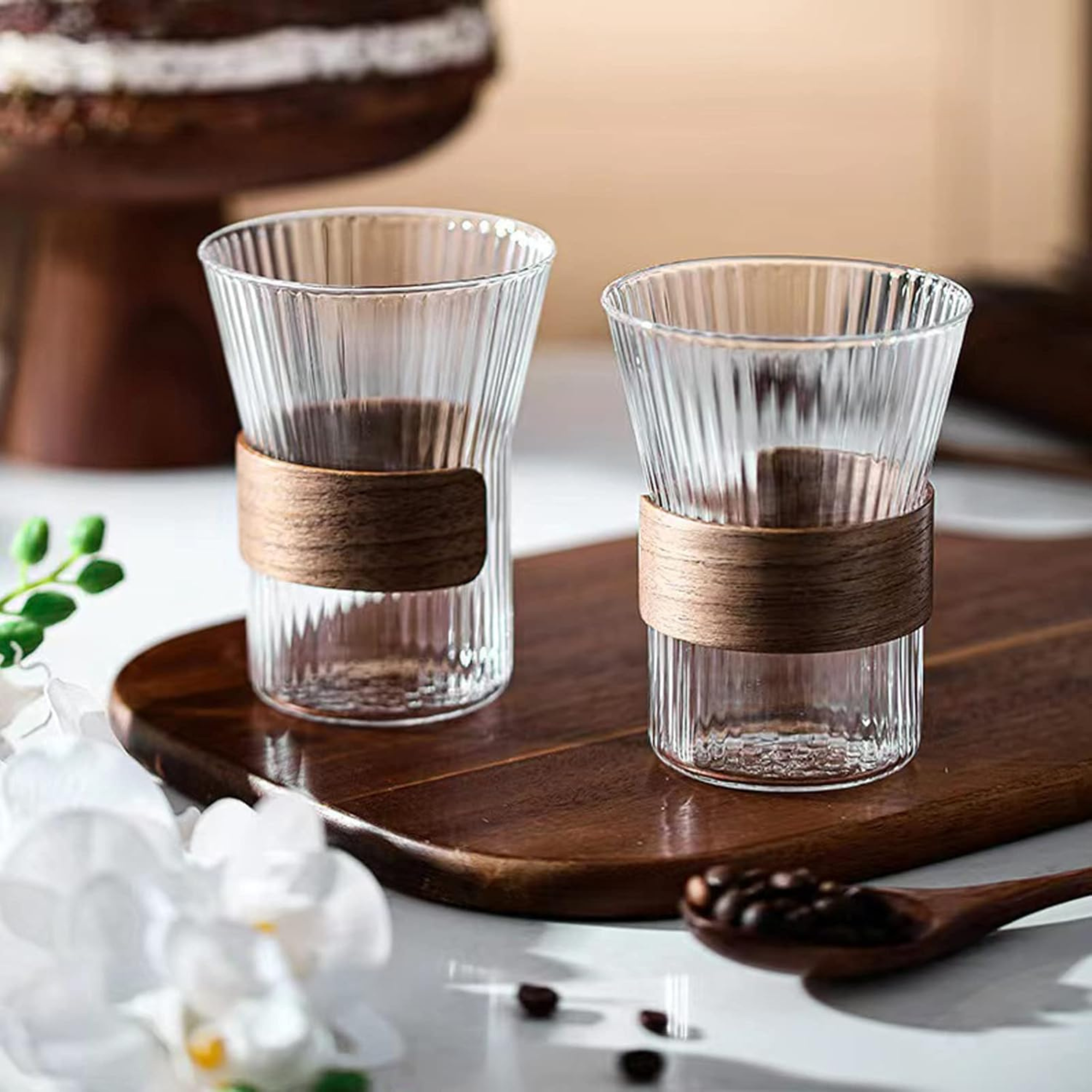 Two clear glass tumblers with wooden bands on a wooden tray, surrounded by coffee beans and flowers.