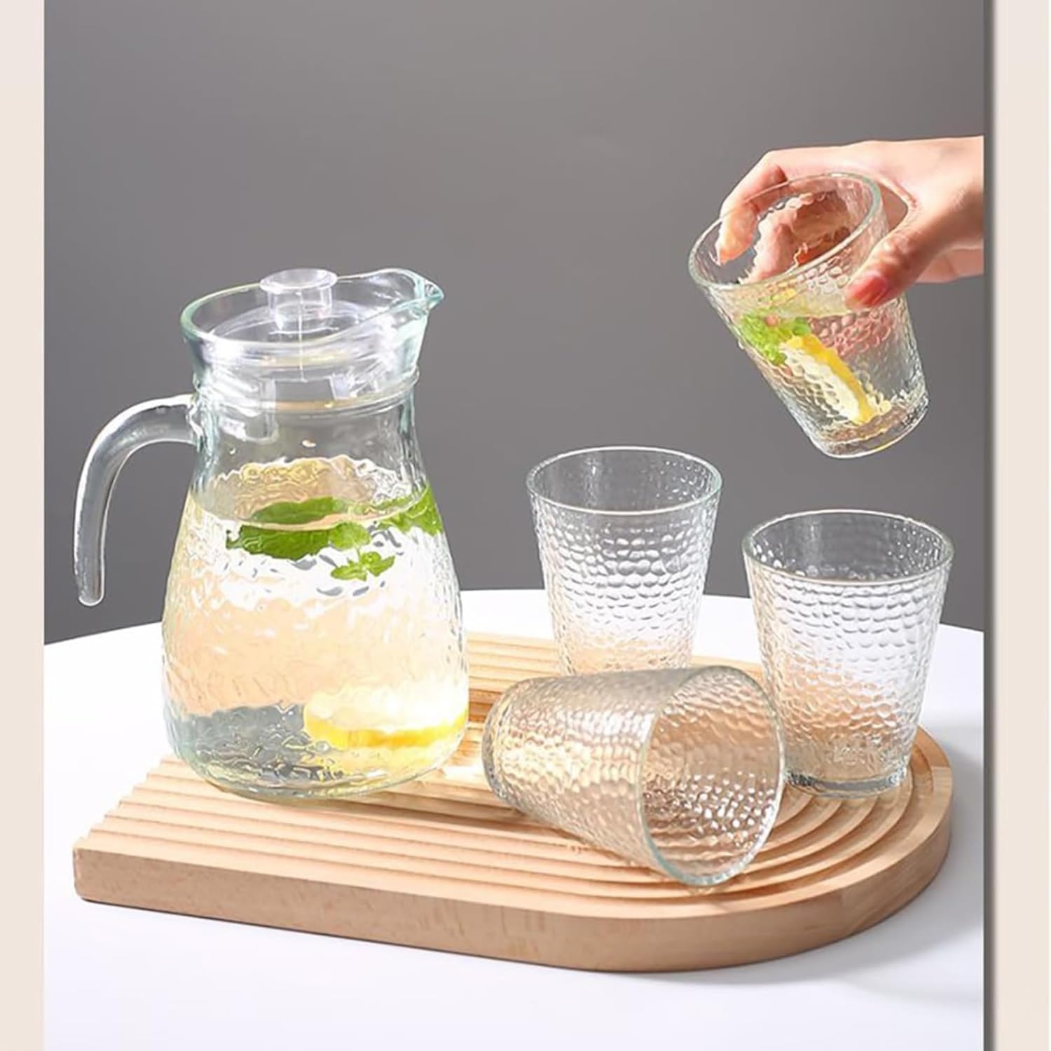 Clear glass pitcher and tumblers on a wooden tray with a hand pouring a drink.