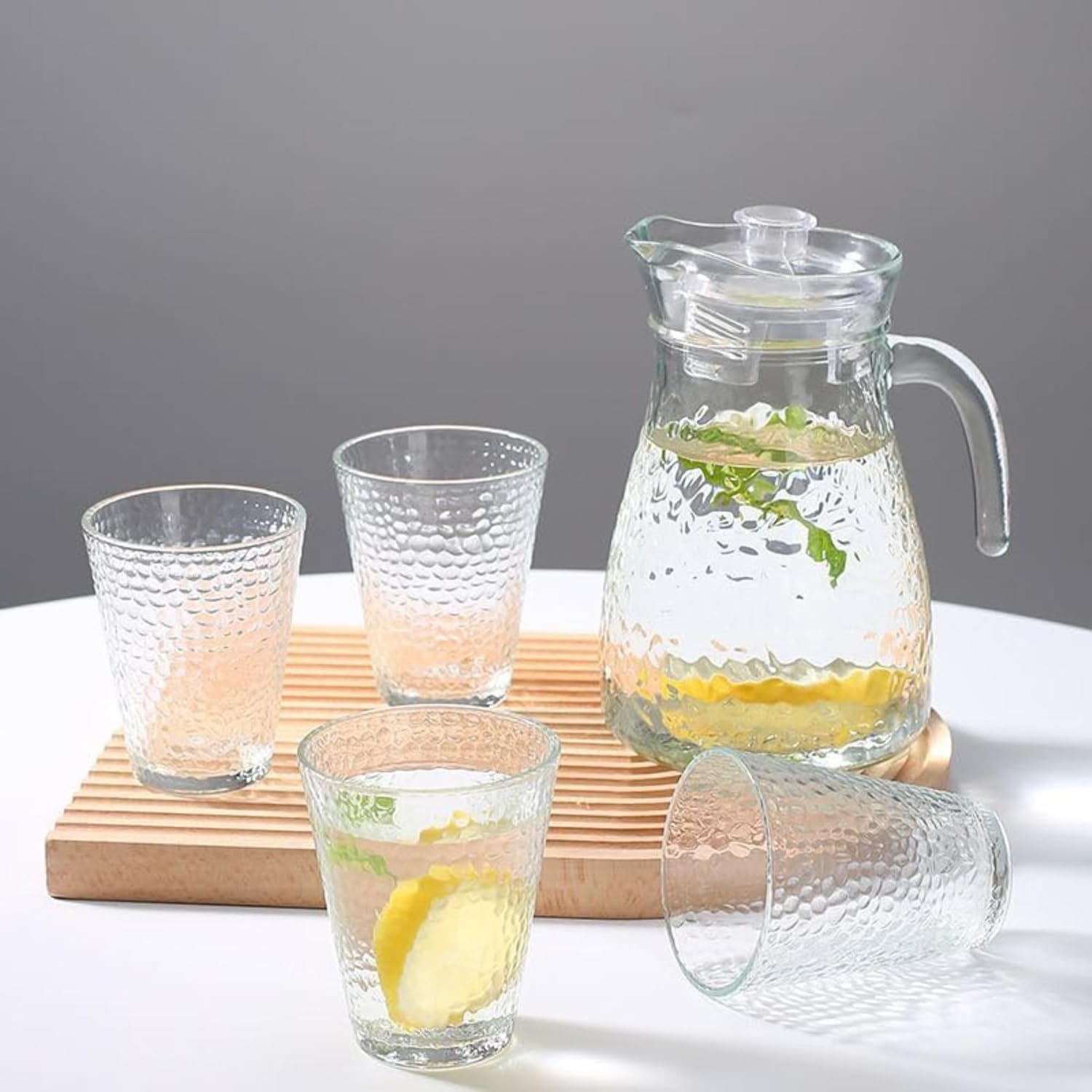 Clear glass pitcher and tumblers with lemon slices on a wooden tray against a gray background