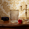 Decorative glass with gold rim on a wooden surface with a red rose and jewelry box, against an ornate wallpaper background.